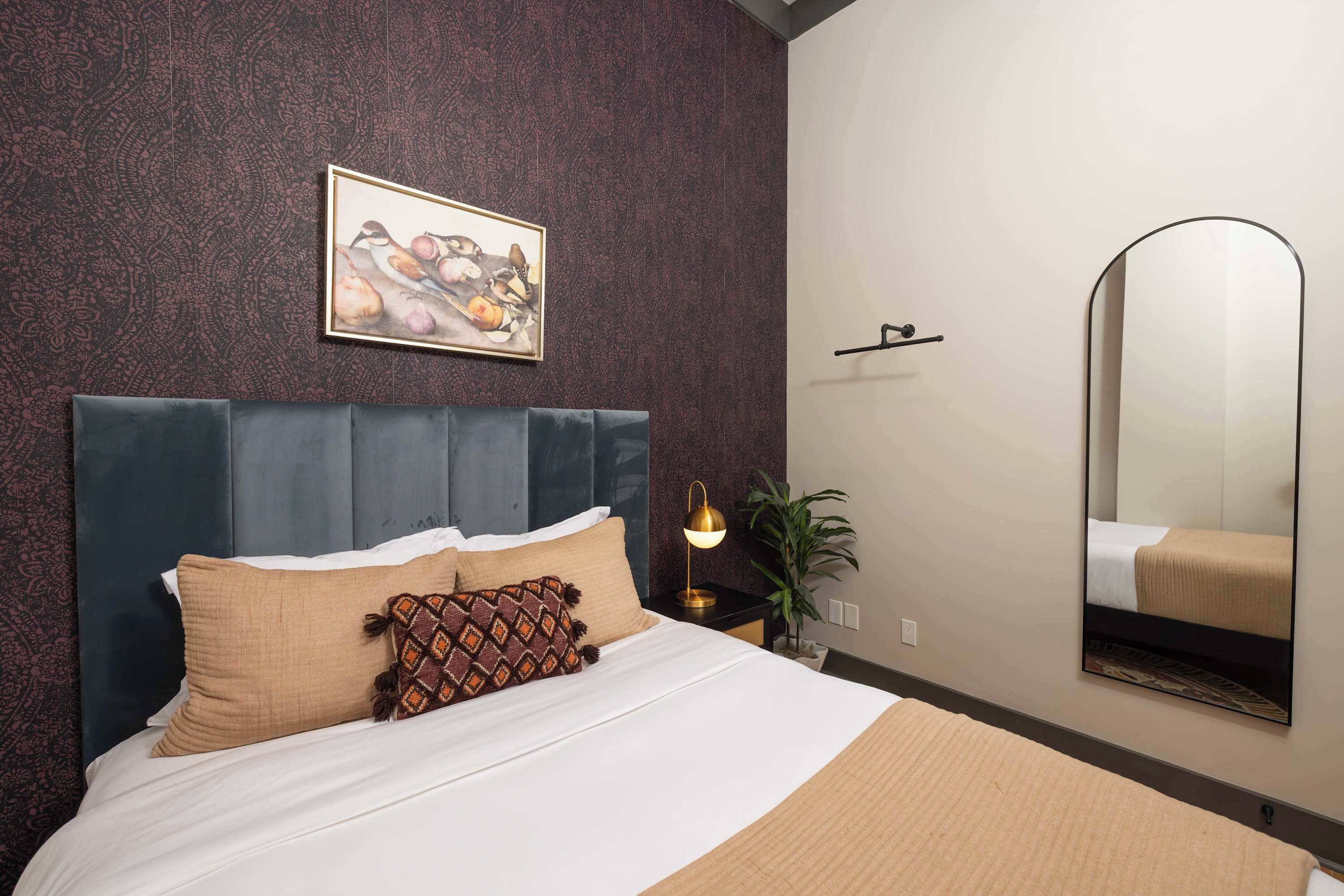 Een hotelkamer met een donker hoofdeinde, een bed met beige beddengoed, een kussen met een patroon, een wandspiegel en een lijst met kunstwerk boven het bed.