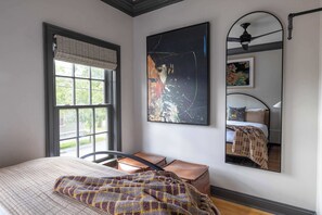 Une chambre avec une grande fenêtre, un lit recouvert d’une couverture à motifs, un tableau abstrait accroché au mur, et un miroir qui reflète le lit et un ventilateur de plafond.