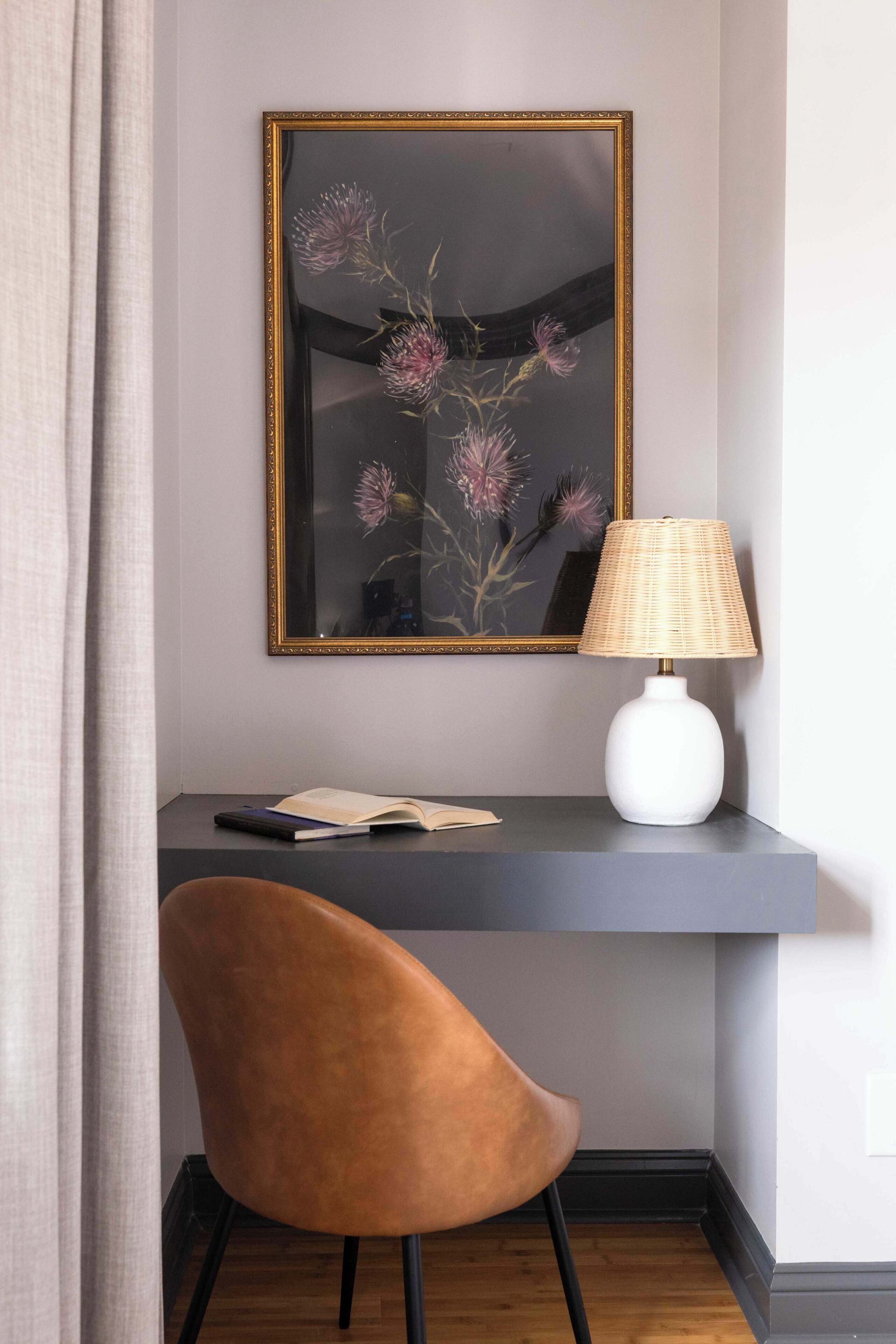 A desk with a lamp, a chair, and a framed artwork on the wall.