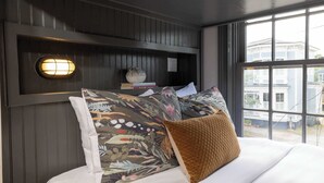 A bedroom with a bed, a nightstand, a wall-mounted lamp, and a view of the outside through a window. - Renovated Townhome Near Forsyth Park | Parking (Savannah)