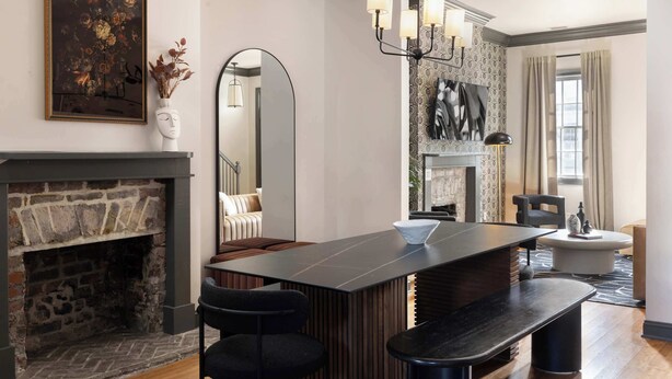 A modern living room with a stone fireplace, a large mirror, a dining area with a long table and chairs, and a chandelier.