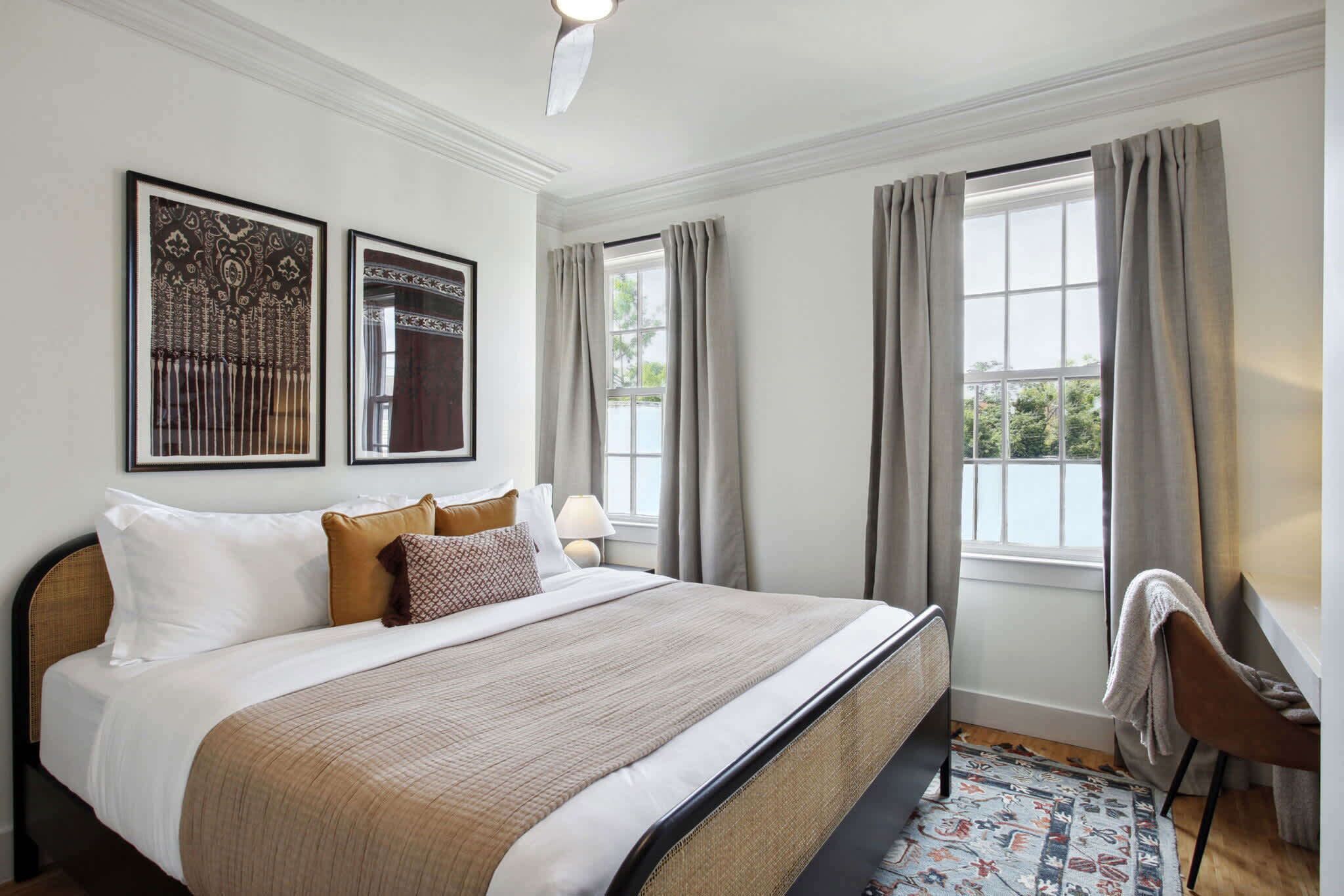 Un dormitorio ordenado con una cama grande, dos cuadros enmarcados, una ventana con cortinas y una silla junto a la ventana.