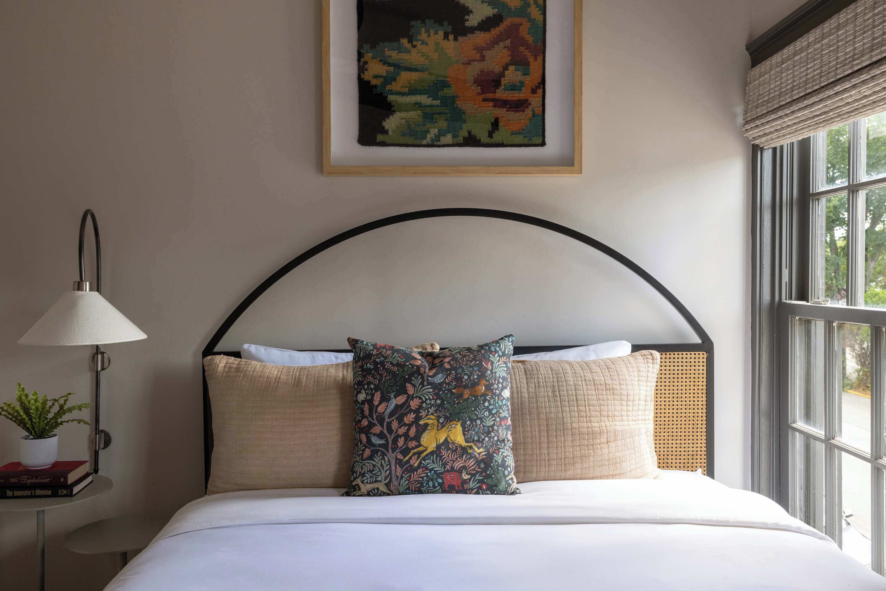 A neatly made bed with a black curved headboard, a colorful floral pillow, and a small table with a lamp and a plant.