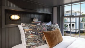 A bedroom with a bed, pillows, a bedside table with books, a wall-mounted light, and a window with a view of a house.