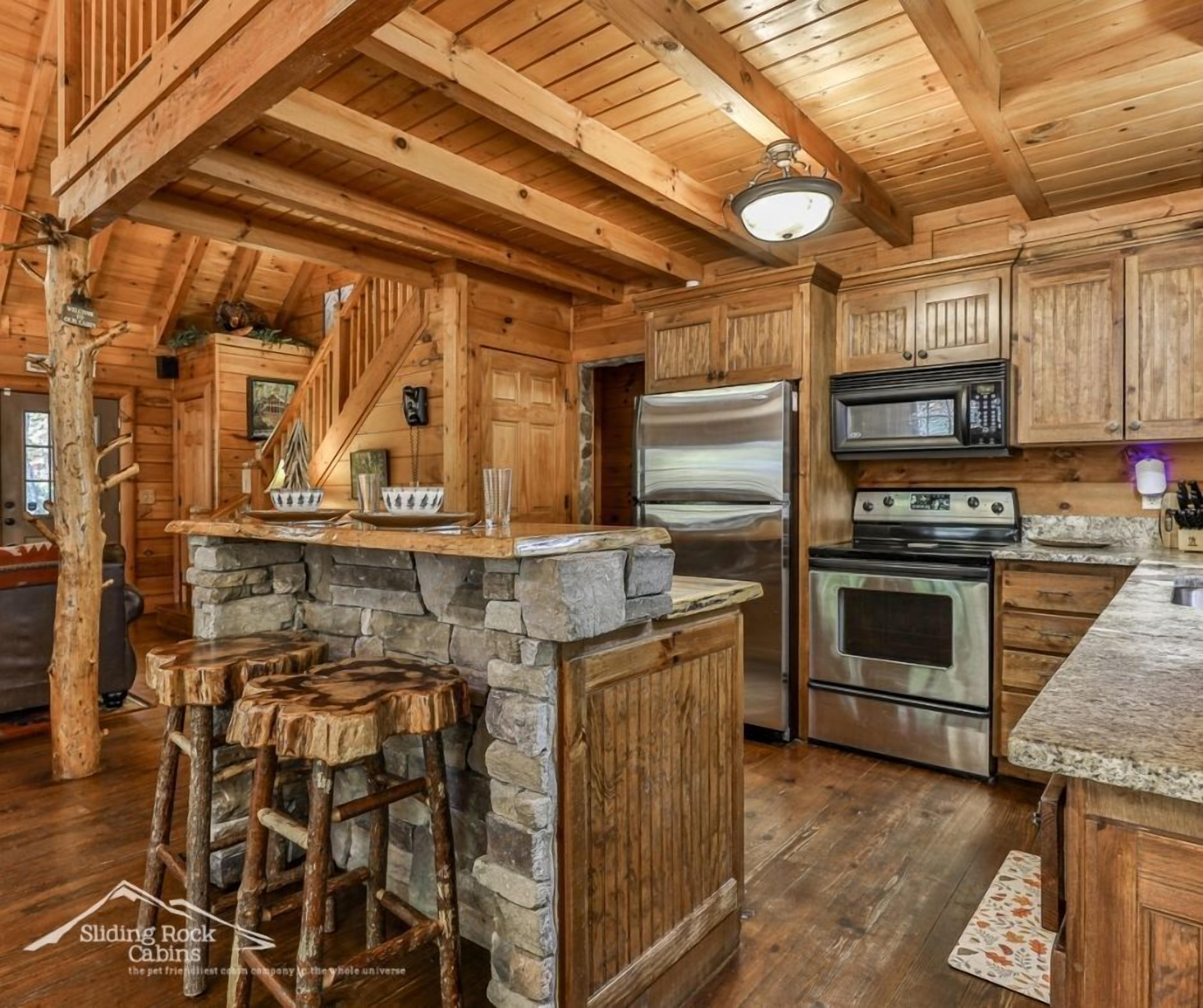 A rustic cabin kitchen with stone countertops, wooden cabinets, a stone fireplace, and a bar stool area.