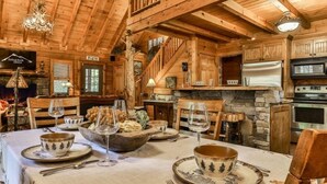 L’intérieur d’un chalet en bois comprend une table à manger pour quatre personnes, des murs en pierre, des poutres en bois, un escalier et un coin cuisine équipé d’appareils électroménagers.