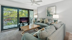 A modern living room with a large window, a ceiling fan, a flat-screen TV, a grey sofa, a wooden coffee table, and two floor lamps. - A-207 - Resort Views and Updated Kitchen! (Naples)
