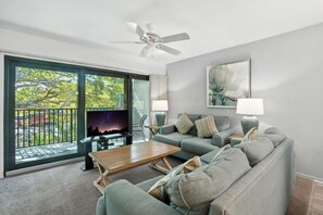 A modern living room with a large window, a ceiling fan, a flat-screen TV, a grey sofa, a wooden coffee table, and two floor lamps.