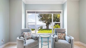 A room with two armchairs, a small round table, and a large window with a view of a tree and water. - Peace River Waterfront Luxury Living - 5-BD w Pool (Port Charlotte)