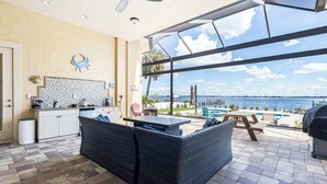 A kitchen with white cabinets, a dining area with wicker furniture, and a large glass door leading to a patio with a view of the sea. - Peace River Waterfront Luxury Living - 5-BD w Pool (Port Charlotte)
