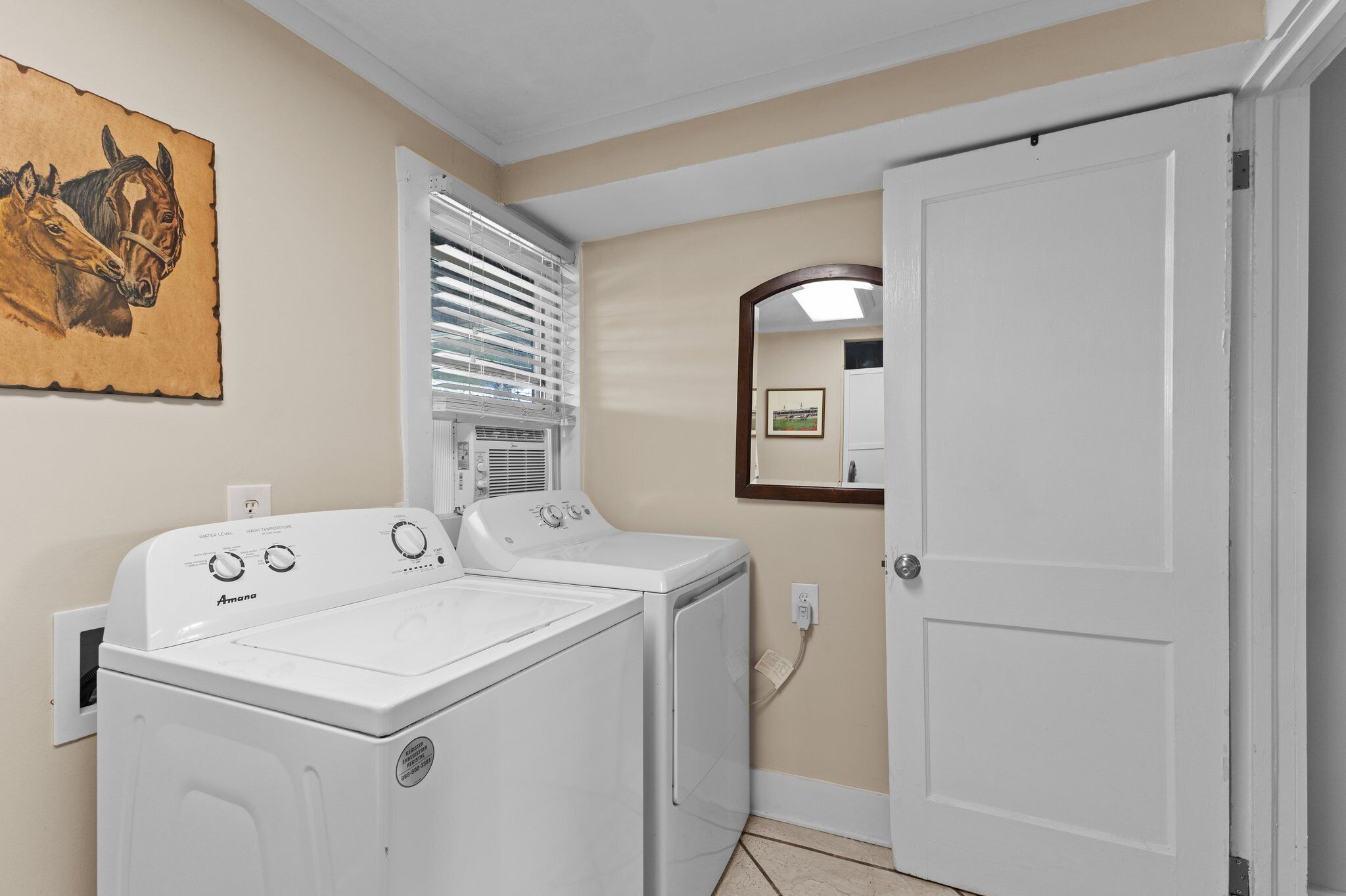 A laundry room with two white front-loading washers and dryers, a window with blinds, a mirror on the wall, and a door.