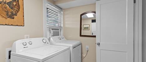A laundry room with two white front-loading washers and dryers, a window with blinds, a mirror on the wall, and a door.