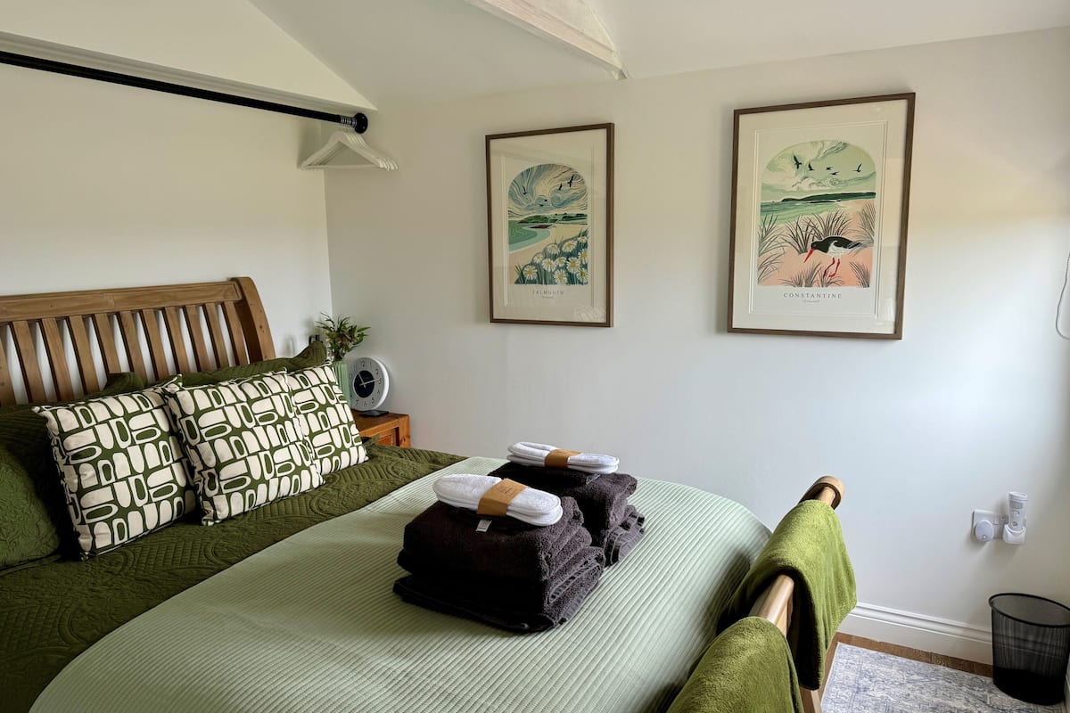 A neatly made bed with green bedding, a wooden headboard, and a bedside table with a clock and a small plant. Two framed botanical prints adorn the wall.