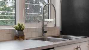 A kitchen with a stainless steel sink, a window with a view of trees, and a potted plant on a wooden stand. - Winston Suite 2BR 1BA - Carriage Suite with Garage (Calgary)