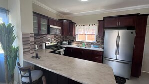A kitchen with dark wooden cabinets, a stainless steel refrigerator, a microwave, and a stove. There are two bar stools and a potted plant. - Charming 2-bedroom townhome with WiFi, AC in beautiful Ocho Rios (Ocho Rios)