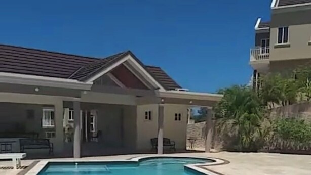A swimming pool with a covered patio area, a house with a red roof, and a hillside with palm trees. - Charming 2-bedroom townhome with WiFi, AC in beautiful Ocho Rios (Ocho Rios)