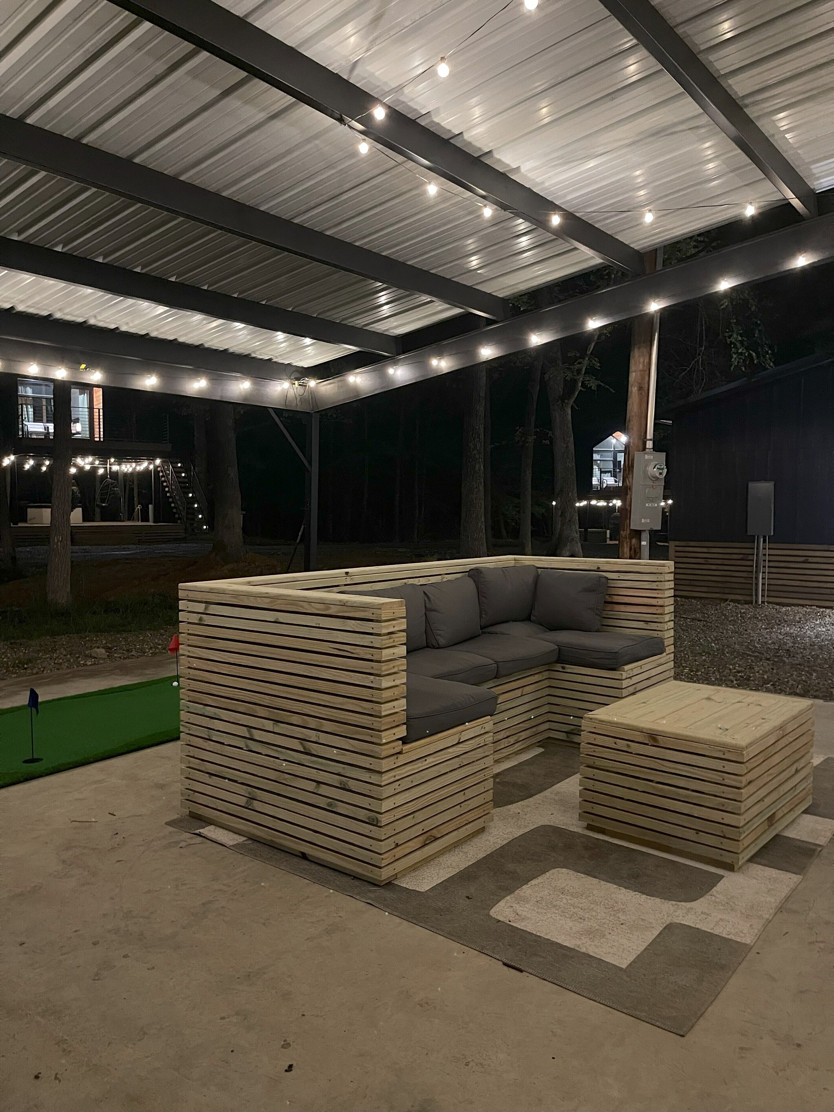 An outdoor seating area with a wooden bench and a sofa, under a covered structure with metal beams and string lights.