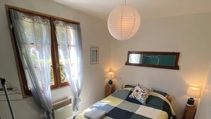 Un dormitorio con una cama, dos lámparas de noche, una lámpara colgante, una ventana con cortinas, un radiador, una silla y una mesita.