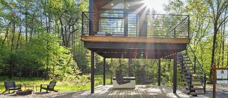 Una cabaña moderna con terraza de madera, asientos al aire libre y una escalera que lleva a un balcón.