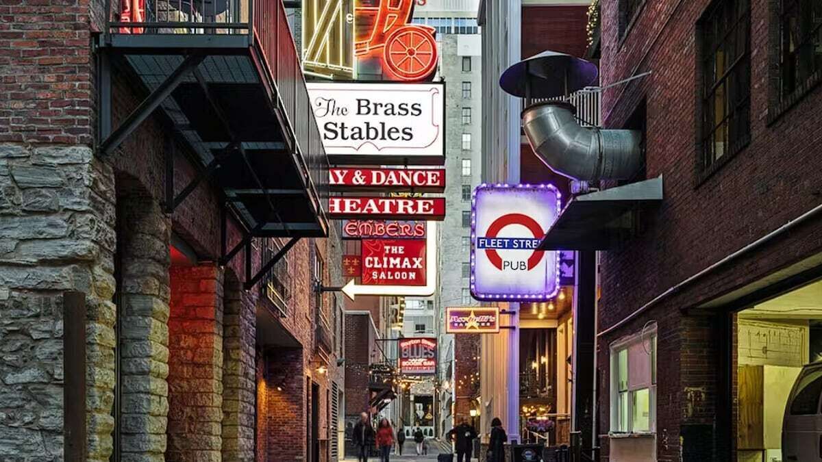 Un callejón estrecho con varios letreros iluminados que anuncian diferentes lugares, como The Brass Stables, un teatro de danza y un pub.