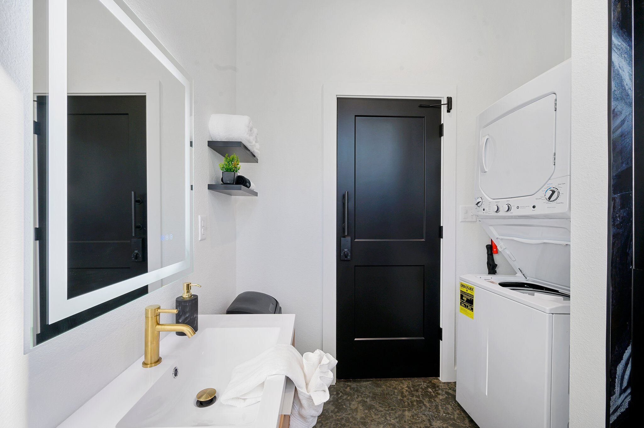 Un baño pequeño con un lavamanos blanco, grifo dorado y un espejo encima. Hay una puerta negra que da a otra habitación, un estante con una planta y una lavadora blanca.