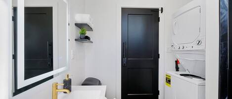 Un baño pequeño con un lavamanos blanco, grifo dorado y un espejo encima. Hay una puerta negra que da a otra habitación, un estante con una planta y una lavadora blanca.