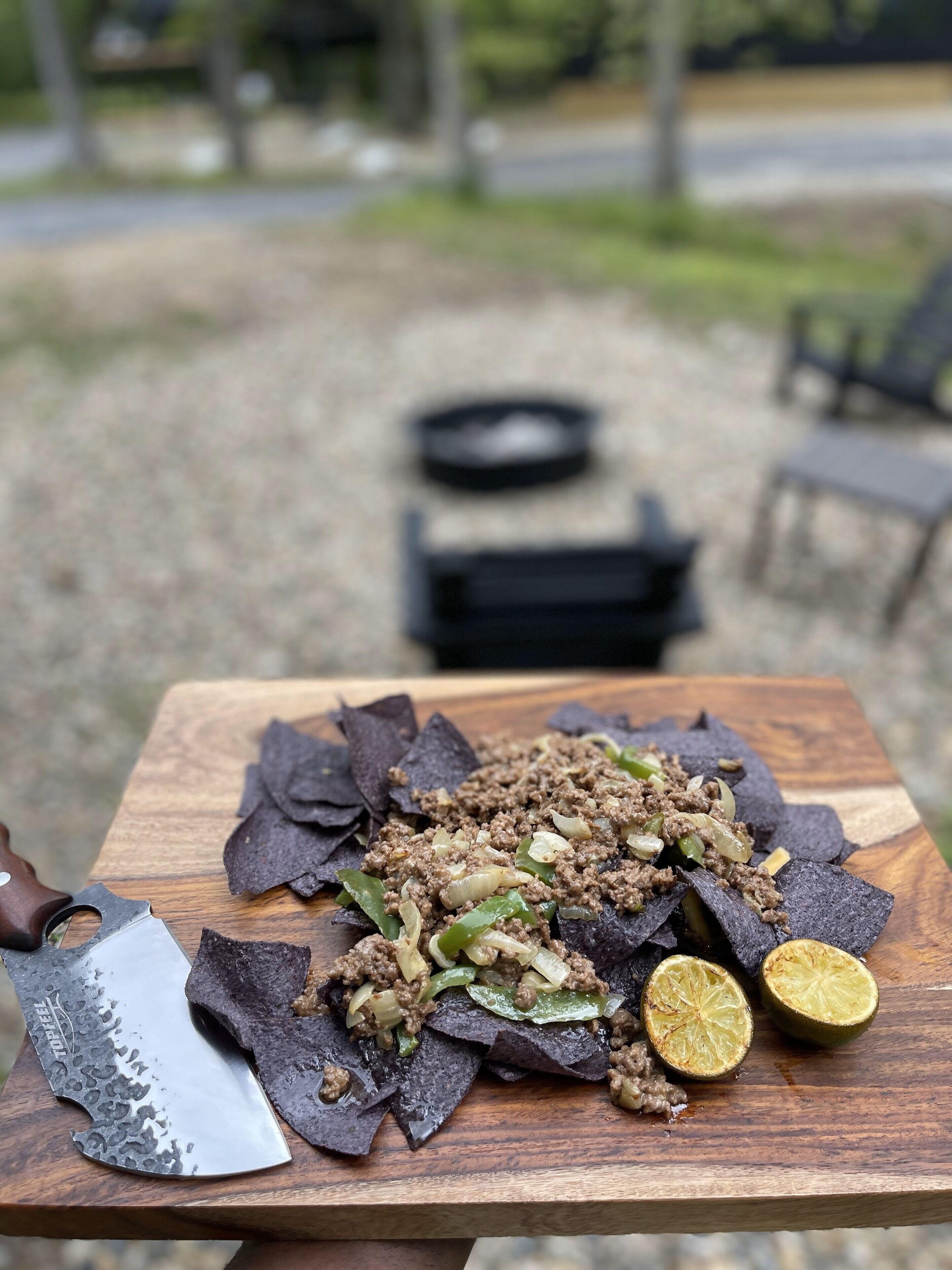 Une table en bois sur laquelle on trouve un plat composé de viande hachée, de poivrons verts et d’oignons, le tout servi sur des chips de tortillas, ainsi qu’un couteau et un demi-citron vert.