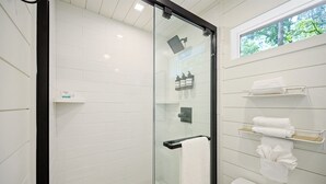 Un baño moderno con cabina de ducha de vidrio, paredes cubiertas de azulejos blancos y ventana con vistas a árboles.