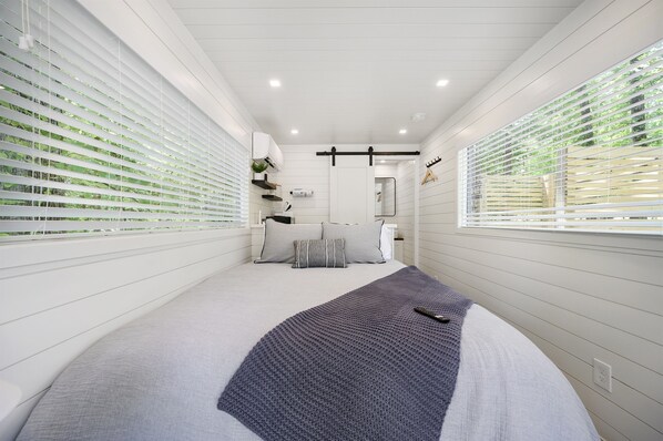 A narrow, white-walled room with a bed, a window, and a door leading to a bathroom.