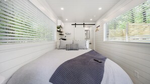 Una habitación pequeña con paredes blancas, una cama, un ventanal y una puerta que da a un baño.