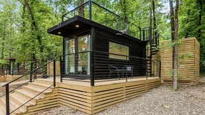Una cabaña moderna con terraza de madera, ventanas de vidrio y una escalera que conduce a una barandilla de metal negro.