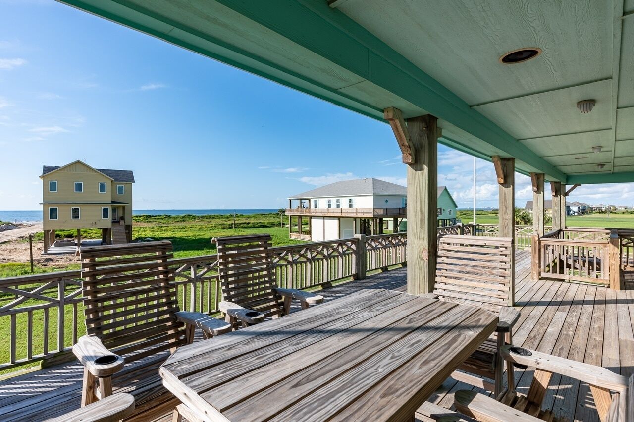 A wooden deck with outdoor seating, overlooking a beach and a house.