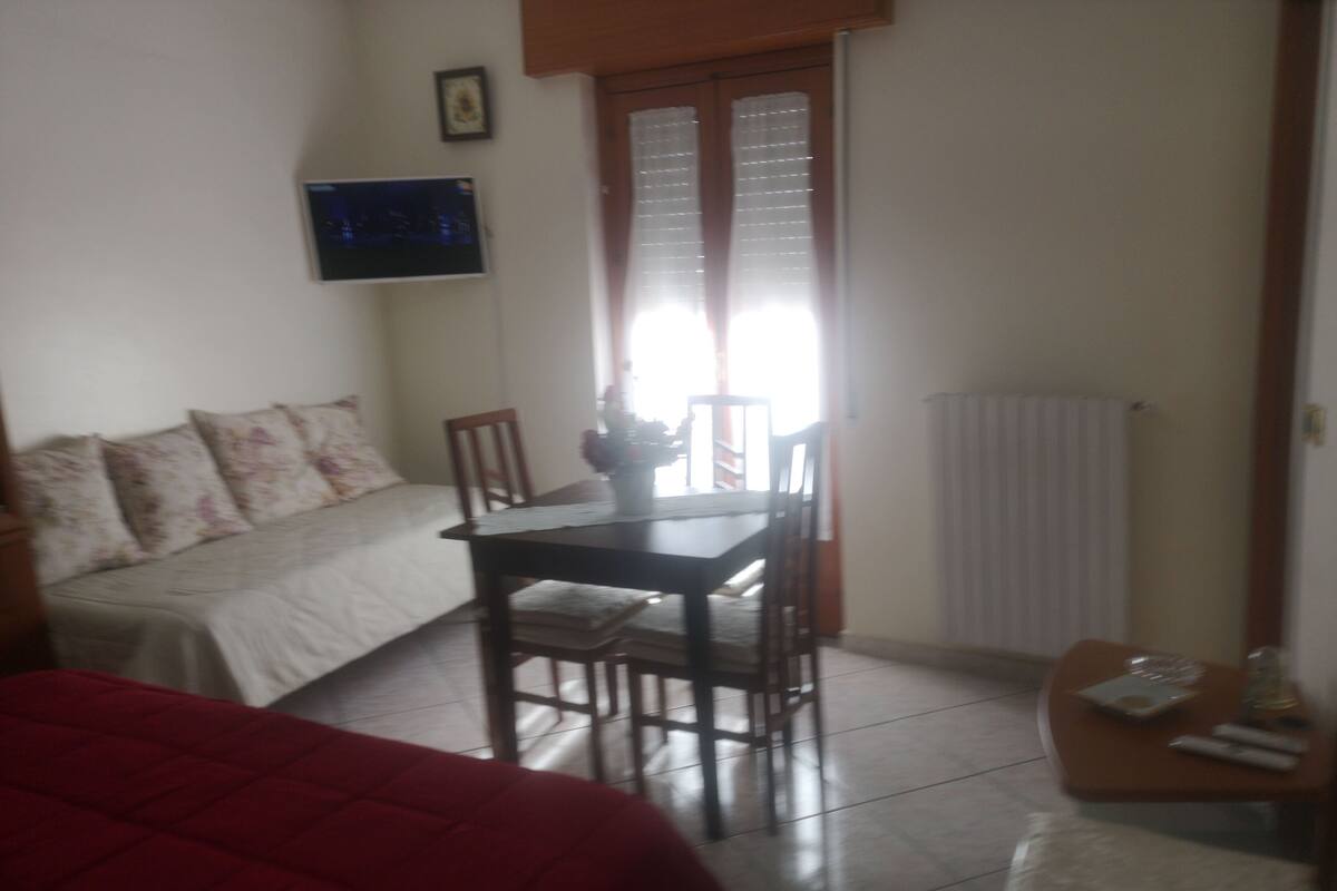 Une pièce comprenant un lit, une table et une chaise. Il y a une fenêtre avec des rideaux, un radiateur et un tableau accroché au mur.