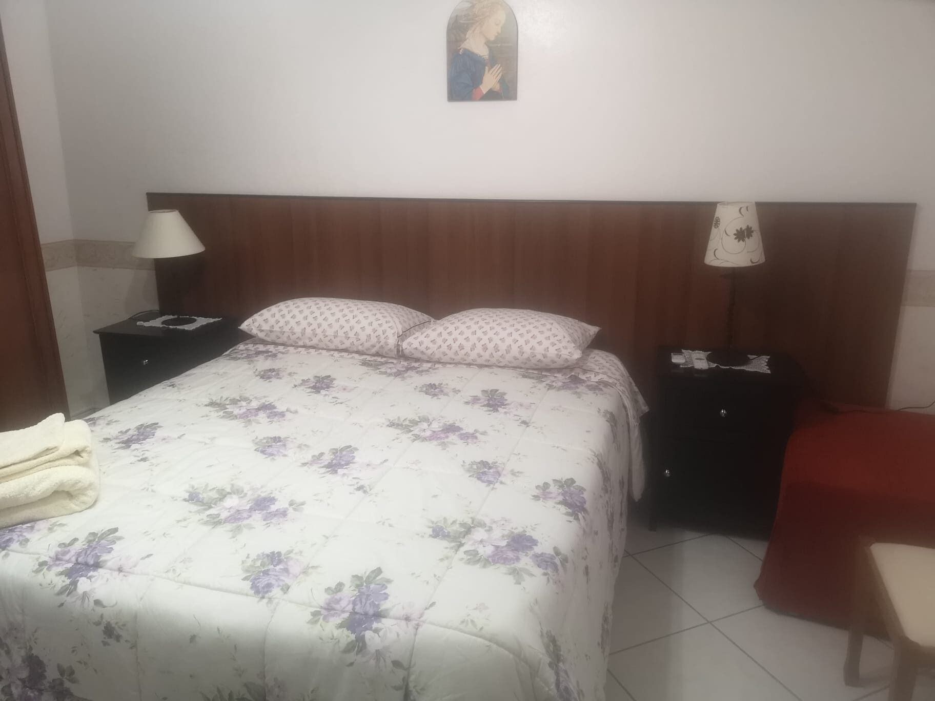 Une chambre à coucher avec une couvre-lit à motifs floraux, deux tables de chevet avec des lampes, une table de nuit, une chaise et une œuvre d’art accrochée au mur.
