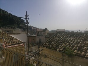 Μια ταράτσα με κεραμίδια, μεταλλική μπαλκονόπορτα και θέα σε μια αστική περιοχή με κτίρια και λόφους στο βάθος.