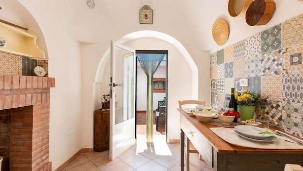 A kitchen with a brick fireplace, a dining table set with plates and cutlery, and a view through an open door to the outside. - 4072 Trull' Itria by Barbarhouse (Martina Franca)