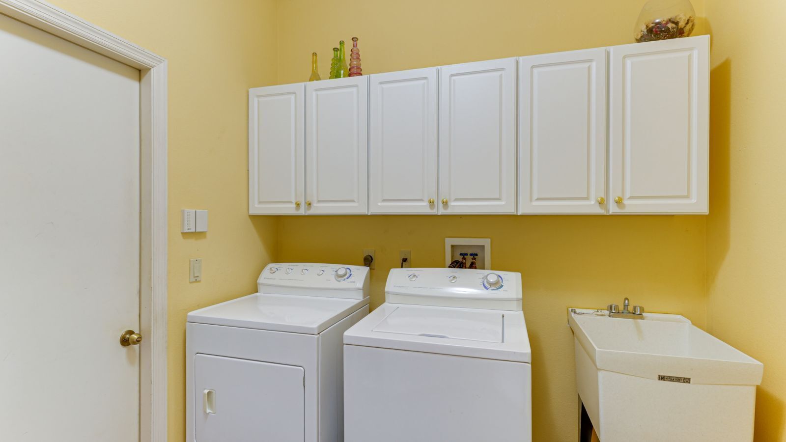 A laundry room with two front-loading washers and a stacked dryer, white cabinetry above, and a white sink.