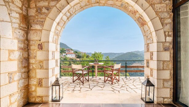 Un arc en pierre mène à une terrasse agrémentée d’une table et de chaises, d’où l’on peut admirer un paysage montagneux.