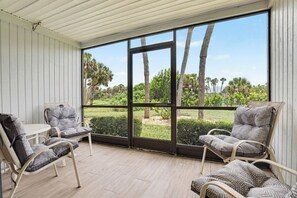 A covered patio with two wicker chairs and a small table, overlooking a well-maintained garden with palm trees and a clear sky. - 2 Bed 2 Bath Condo on Golf Course in Quiet Safe Community, 4 Miles to Beach (Venice)