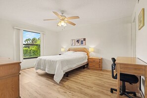 Ein Schlafzimmer mit einem Bett, Nachttischen aus Holz, einem Schreibtisch mit Stuhl, einem Deckenventilator und einem Fenster mit Blick auf Grünflächen.