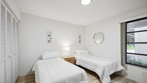 A bedroom with two beds, a bedside table with a lamp, a round mirror, and a window with a view of greenery. - 2 Bed 2 Bath Condo on Golf Course in Quiet Safe Community, 4 Miles to Beach (Venice)