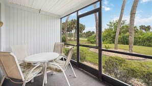A covered patio with a round table and chairs, large windows offering a view of palm trees and greenery. - 2 Bed 2 Bath Condo on Golf Course in Quiet Safe Community, 4 Miles to Beach (Venice)
