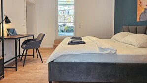 Ein modernes Schlafzimmer mit einem Bett, einem Schreibtisch mit Laptop, einem Stuhl und einem Fenster mit Blick auf Gebäude.