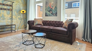Ein Wohnzimmer mit einem braunen Sofa, zwei runden Tischen, einer Stehlampe und einem großen Fenster mit Blick auf Gebäude.