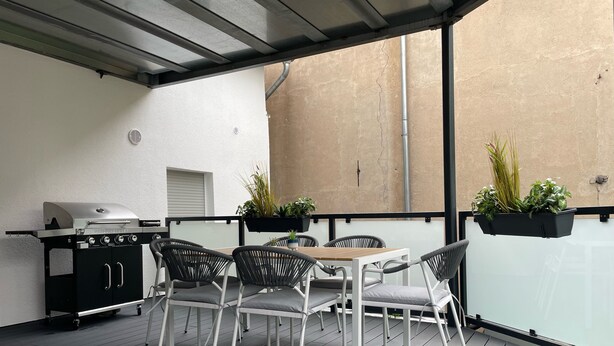 Eine Außenpatio mit Grill, weißen Korbstühlen, einem Tisch und Topfpflanzen.