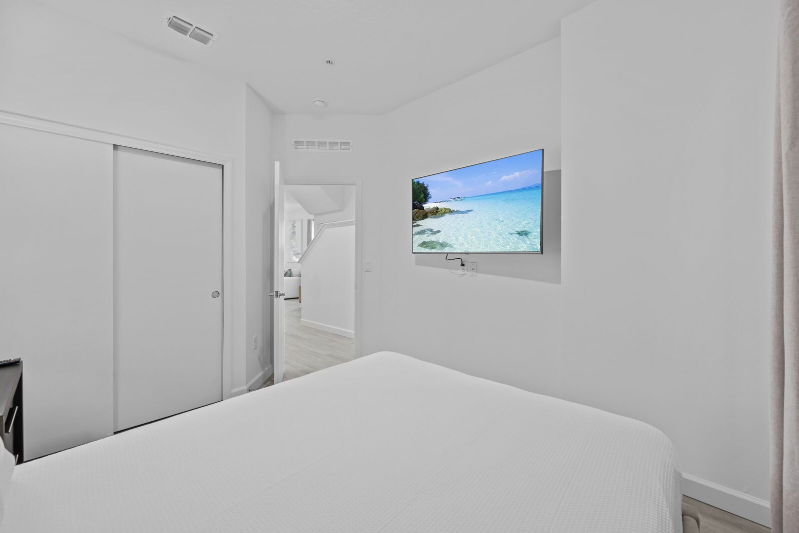 A hotel room with a bed, a TV mounted on the wall displaying a beach scene, and a door leading to another room.