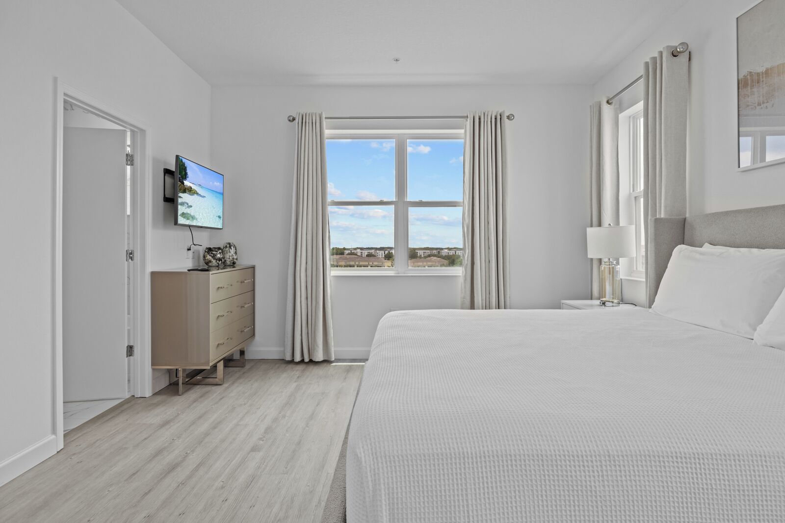 Une chambre moderne avec un grand lit, un téléviseur fixé au mur, une commode et une fenêtre avec des rideaux.
