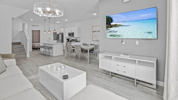 Un salon moderne avec un canapé blanc, une table basse, un téléviseur fixé au mur et un coin repas avec une cuisine en arrière-plan.