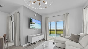 Un salon moderne doté d’un téléviseur à écran plat, d’un canapé blanc et d’une grande fenêtre donnant sur une piscine et un espace verdoyant.