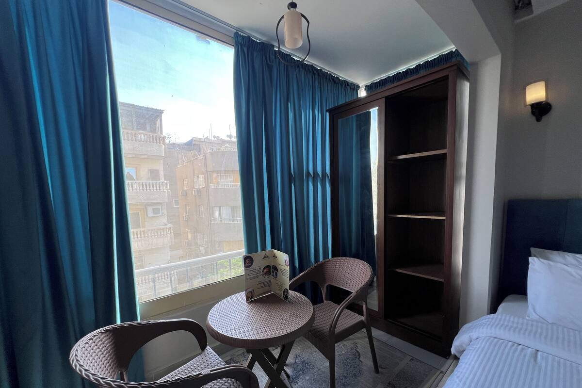 A hotel room with a large window, a bed, a small table with two chairs, and a bookshelf.